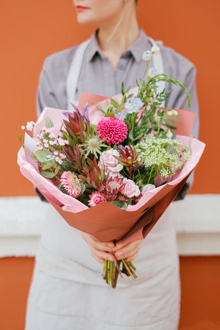 A person dressed in a light grey shirt and white apron holds a vibrant bouquet of fresh flowers wrapped in pink and peach-colored paper, positioned against a warm orange background. The bouquet featur