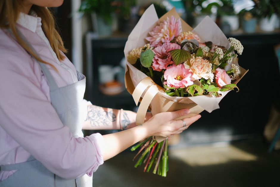 A person with long, light brown hair, wearing a light pink shirt and a white apron, holding a bouquet of fresh flowers. The bouquet includes pink peonies, soft peach carnations, white baby's breath, a