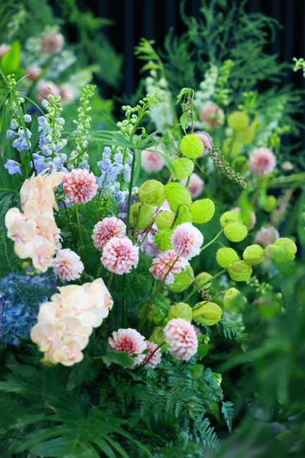 A lush bouquet featuring delicate pink and peach carnations, vibrant green billy balls, light purple waxflowers, and various shades of green foliage, including ferns and leafy stems. The flowers are a