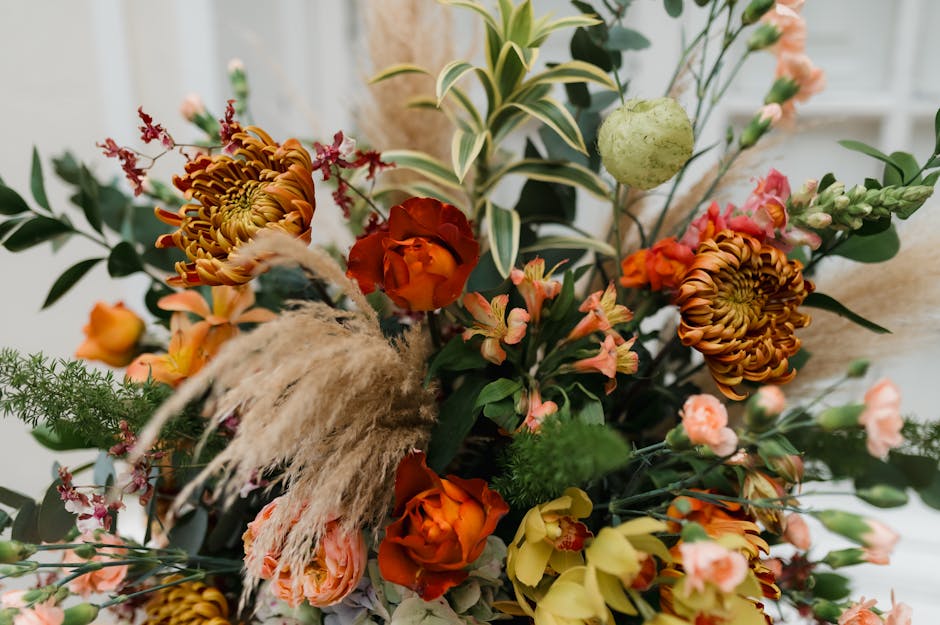 A vibrant floral arrangement featuring a mix of orange roses, brown and yellow chrysanthemums, soft pink miniature roses, and delicate pink and peach blossoms. The bouquet is complemented by greenery 