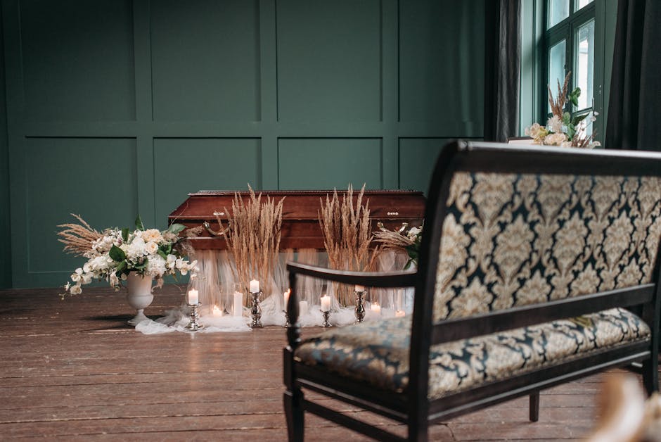 A wooden coffin is adorned with floral arrangements featuring white roses, eucalyptus, and dried pampas grass, placed on a white cloth with small lit candles surrounding it. To the left, a white vase 
